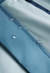 Vandtæt stof i lys blå og blågrøn farve med dråber på overfladen, der fremhæver en glat tekstur og detaljer ved syningerne.