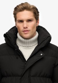 Jonge man met gestileerd lichtbruin haar, gekleed in een zwart gewatteerd jack en een lichtgrijze geribde turtleneck trui tegen een witte achtergrond.
