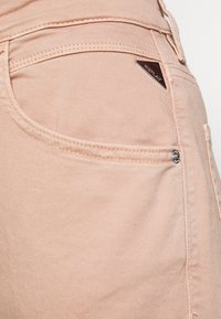 Vaaleanpunaiset farkut, joissa on sileä pinta, pienen metallilogon yksityiskohta ja ompelu yksityiskohtia takataskun lähellä.