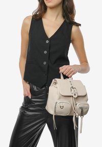 Beżowy plecak skórzany z klapą, srebrnymi elementami i przednimi kieszeniami. Modelka ma na sobie czarną bezrękawnik i skórzane spodnie.