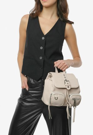 Zaino in pelle beige con patta, accessori in argento e tasche frontali. Il modello indossa un gilet nero senza maniche e pantaloni di pelle.