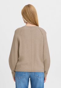Pull en tricot marron clair avec un col rond, des manches raglan et des coutures discrètes. Associé à un jean bleu clair.