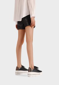 Osoba ubrana w czarne skórzane szorty, białą koszulę z długim rękawem oraz czarne buty sportowe na platformie z białymi podeszwami, stojąca na jednolitym tle.