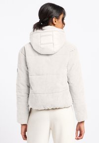 Lichtgrijze corduroyjas met een pluchen witte capuchon, ribbeltextuur en elastische tailleband. Heeft een opvallend stikselpatroon.