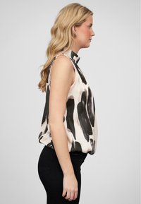 Blouse sans manches en crème avec de grands motifs abstraits noirs, présentant un col montant et une coupe ample. Associée à un pantalon noir.