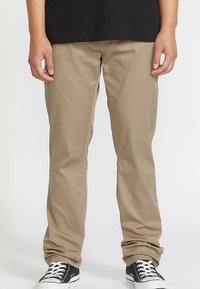 Pantalon en coton beige à coupe droite, porté avec des baskets noires et blanches. Le tissu semble lisse, sans motifs ni ornements visibles.