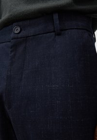 Pantaloni da abito blu navy in tessuto strutturato, con chiusura frontale a bottone e una leggera lucentezza. È visibile un motivo sottile e intricato.