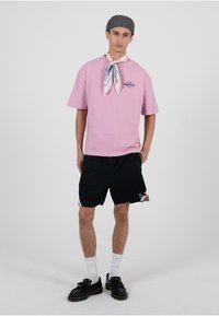 Roosa lühikeste varrukatega t-särk tekstiga "KEELATUD", mustade lühikeste pükstega, valgete keskpikkade sokkidega ja mustade kingadega. Aksessuaarideks on mustriga sall ja hall nokamüts.