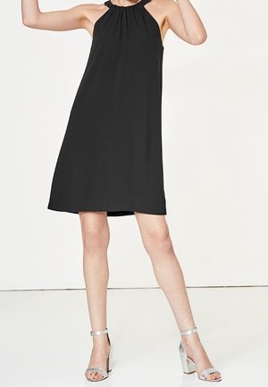 Vestito nero senza maniche con scollo arricciato, silhouette fluida e lunghezza sopra il ginocchio, abbinato a sandali con tacco argentati.