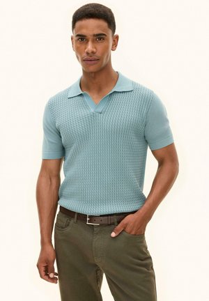 Mand iført lyseblå tekstureret kortærmet poloshirt, stoppet ned i olivengrønne bukser med brunt bælte, stående mod en ensfarvet baggrund.