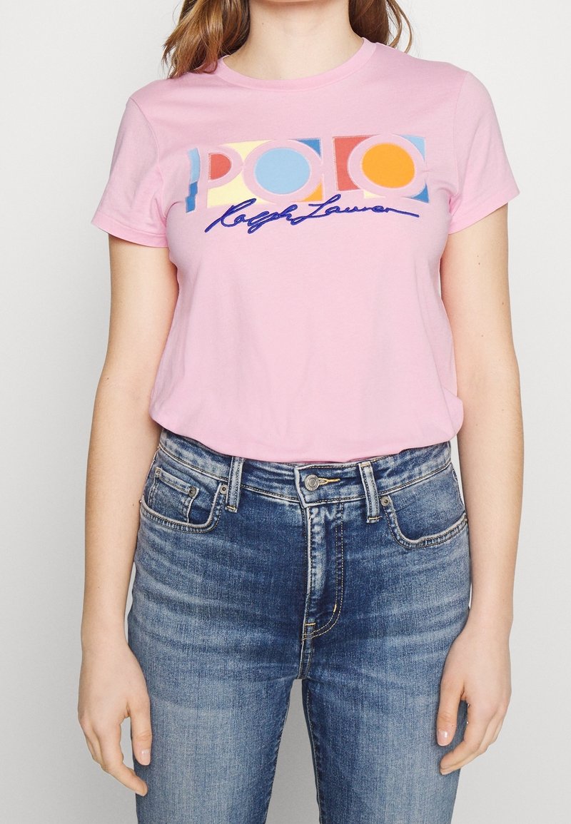 Žena oblečená v růžovém tričku s krátkým rukávem s barevným nápisem „POLO Ralph Lauren“, doplněné modrými džínami s vysokým pasem.