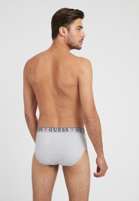 Graue Baumwollunterwäsche mit passgenauer Form. Verfügt über einen navyblauen Bund mit "GUESS"-Schriftzug in Weiß. Glatte Textur, minimalistischem Stil.