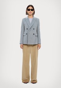 Blazer double boutonnage en velours côtelé bleu clair avec des boutons noirs, associé à un pantalon large beige. Le mannequin porte des lunettes de soleil noires et des boucles d'oreilles.