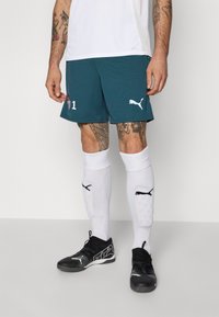 Teal sportshorts met witte accenten, voorzien van logo en nummer. Zwarte schoenen met gestructureerde zolen, gecombineerd met witte scheenbeschermers.
