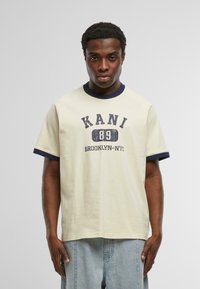 Krémové tričko s námořnicky modrým žebrovaným límcem a rukávy. Na přední straně je potisk: "KANI 89 BROOKLYN-NYC" v tučném, opotřebovaném písmu.