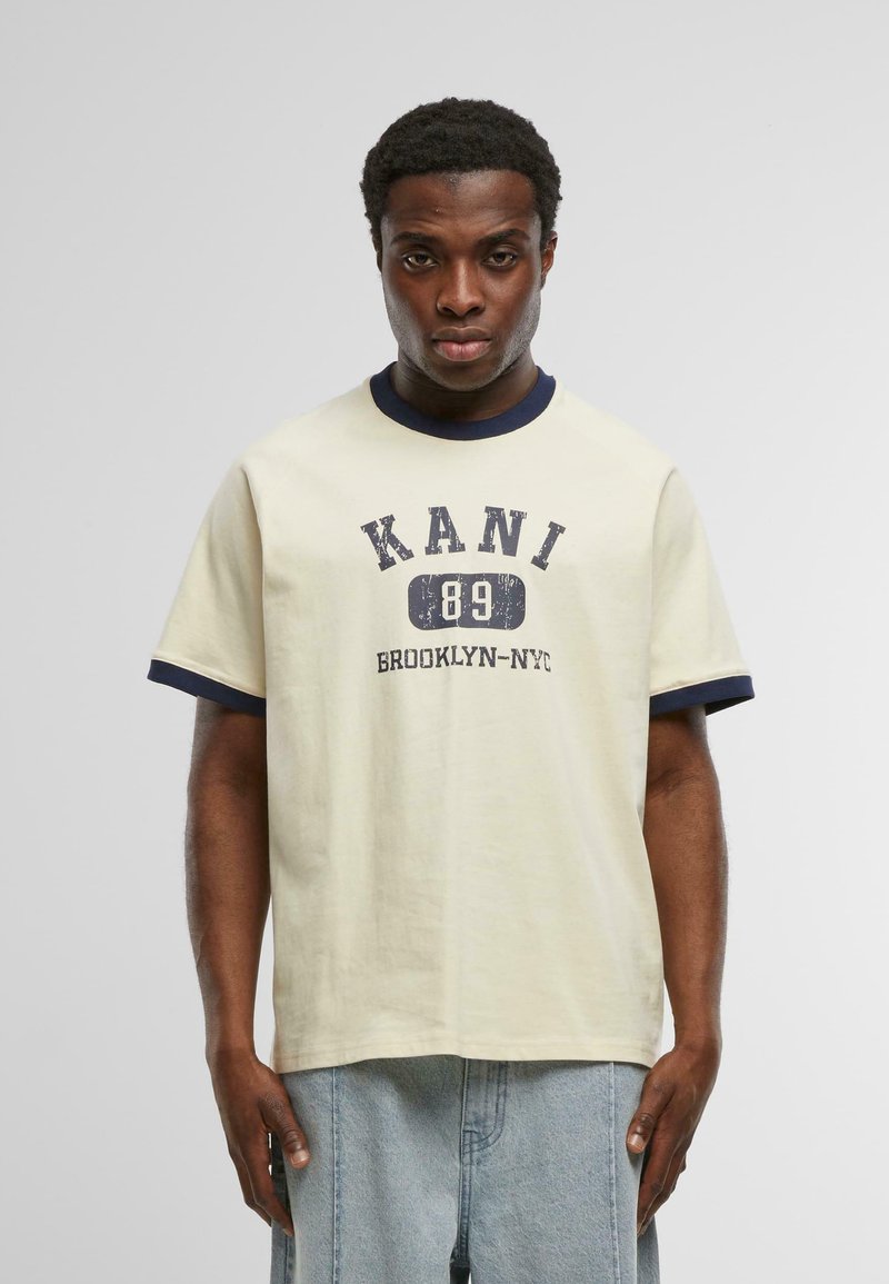 Krémové tričko s námořnicky modrým žebrovaným límcem a rukávy. Na přední straně je potisk: "KANI 89 BROOKLYN-NYC" v tučném, opotřebovaném písmu.