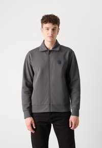 Giacca grigia con zip e colletto alto, realizzata in tessuto liscio. Presenta un logo esagonale sul petto e polsini a coste.