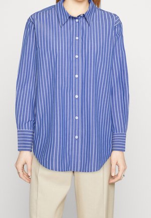 Persoon draagt een blauw overhemd met lange mouwen en witte verticale strepen, gecombineerd met beige broek, staand tegen een effen achtergrond.