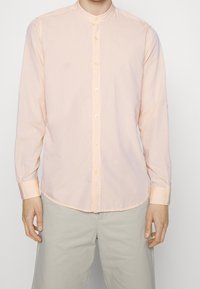 Light peach long-sleeve shirt with a stand-up collar, featuring a subtle pattern and light-colored buttons. Worn with light gray pants.