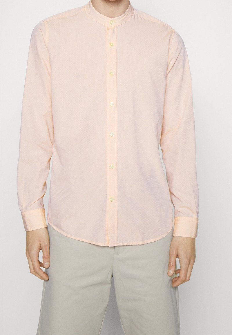 Light peach long-sleeve shirt with a stand-up collar, featuring a subtle pattern and light-colored buttons. Worn with light gray pants.