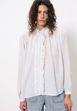 Weiße transparente Bluse mit vertikalen blauen Streifen, gerafftem Vorderplacket, langen Ärmeln und klassischem Kragen. Kombiniert mit Denim-Shorts.