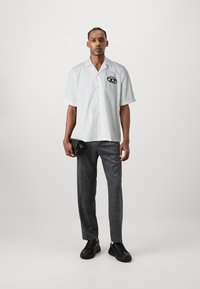 Kortærmet hvid skjorte med et sort logo, kombineret med skræddersyede grå bukser. Outfit inkluderer en sort clutch og sorte sneakers.