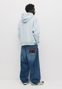 Felpa leggera blu con un taglio rilassato, abbinata a jeans blu oversize con una grafica rossa e nera sulla tasca posteriore, e sneakers nere.