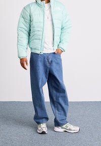 Ljus turkos puffjacka, vit T-shirt, lösa blå jeans och grågröna löparskor. Jackan har en hög krage och dragkedjefickor.
