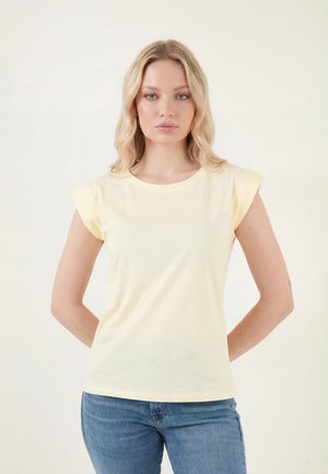 Mujer rubia de pie con los brazos detrás de la espalda, vestida con una camiseta sin mangas amarillo pálido y vaqueros azules sobre un fondo liso y claro.