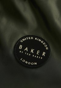 Parche circular de tela negra con letras doradas en relieve. El texto incluye "BAKER", "by TED BAKER", "LONDRES" y "REINO UNIDO". Fondo mate.