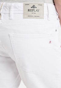 Vita denimjeans med en läderetikett som visar "REPLAY" på baksidan, med två bakfickor och en subtil röd sömnadsdetalj.