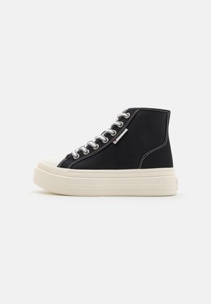 Zwarte hoge sneaker met dikke witte plateauzool, witte veters en contrasterende witte stiksels op een effen lichte achtergrond.