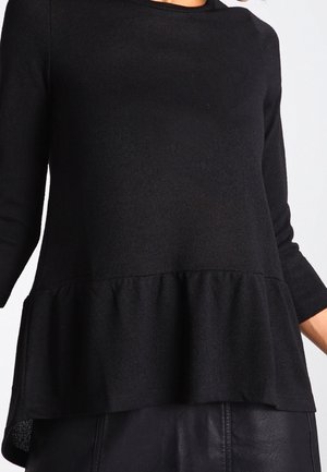 Musta pitkähihaisen puseron, jossa on röyhelöhelma, ja käytetty mustun hameen tai housujen päällä, esitetty olkapäiltä lantioihin asti.