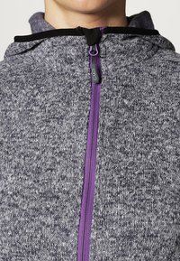 Szary rozpinany hoodie z purpurowym zamkiem błyskawicznym i czarnymi akcentami. Teksturowana tkanina przedstawia nakrapiany wzór, a kaptur jest zintegrowany z designem.