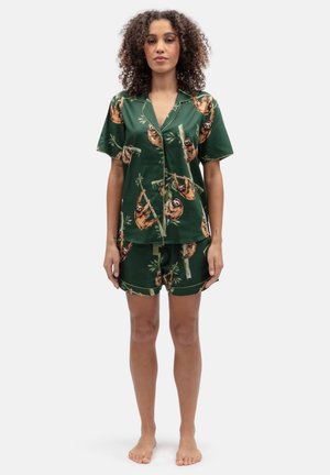 Vrouw staat blootsvoets en draagt een donkergroen kort mouwen overhemd met knopen en shorts met luiaard- en bamboeprint.