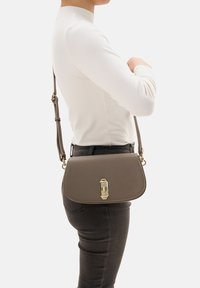 Braune Leder-Crossbody-Tasche mit einem goldenen Verschluss, halbstrukturierter Form und verstellbarem Riemen, getragen über einem weißen Langarmoberteil.