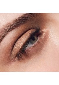 Close-up van een oog met glinsterende bronzen oogschaduw, donkere wimpers en een lichtgekleurde iris. De huid lijkt glad met een natuurlijke glans.