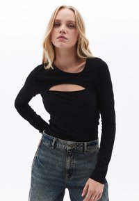 Schwarzes Langarmoberteil mit einem Ausschnitt vorne, aus glattem Stoff. Kombiniert mit hoch taillierten blauen Denim-Jeans im klassischen Design.