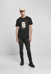 Fekete pamut póló, amelyen elöl kezek nyomtatott mintázata található, fekete slim fit farmernadrággal és fekete sportcipővel párosítva. Beige sapka is tartozik hozzá.