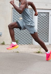 Un athlète portant un débardeur gris et un short gris court en baskets Nike roses vives et jaunes. En arrière-plan, un mur texturé et un motif en grille.