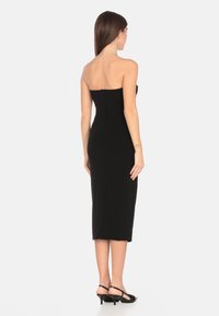 Un abito midi nero, aderente e senza spalline, con una texture liscia e una chiusura con zip sulla schiena, abbinato a sandali neri con tacco.