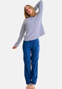 Gestreept wit en marineblauw langemouwen topje, gecombineerd met effen blauwe broek met trekkoord. Het model staat blote voeten en poseert met een opgestoken arm.