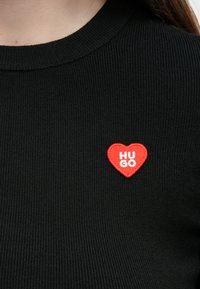 Camisola preta canelada com gola redonda. Apresenta um remendo em forma de coração vermelho com o texto "HUGO". Design simples e ajustado.