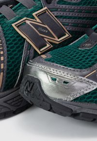 Chaussures de course en mesh vert avec embout métallique gris, semelle en caoutchouc et accents dorés, présentant un logo "N" bien en vue.