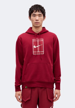 Giovane uomo che indossa una felpa con cappuccio rossa Nike con logo bianco e pantaloni cargo rossi abbinati su uno sfondo grigio uniforme.