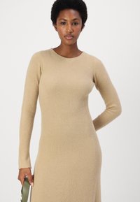 Abito maglione beige a coste con maniche lunghe e scollatura rotonda. Presenta una silhouette aderente e un tessuto morbido e testurizzato.