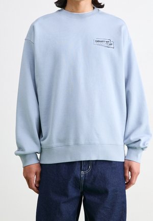 Osoba oblečená v světle modré mikině Carhartt WIP s logem na hrudi, v kombinaci s tmavě modrými džínami, stojící před bílým pozadím.