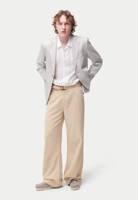 Blazer gris clair, chemise blanche à boutons, pantalon beige à jambes larges, et chaussures grises enfilables avec une semelle texturée. Look simple et ajusté.