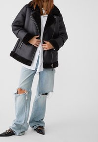 Person som bär en överdimensionerad svart fårskinnsjacka, vit skorta, trasiga ljusblå jeans och svarta spetsiga skor med remmar.