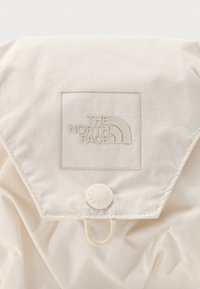 Μπεζ υφασμάτινο τμήμα ενός σακιδίου με καπάκι, κουμπί με πατιλέτα και ανάγλυφο λογότυπο "The North Face".